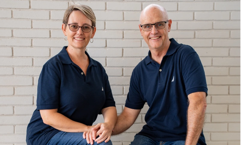 YDL Homes - Derek and Lee sitting on a bench with a railway tile wall background, both in blue polo shirts.